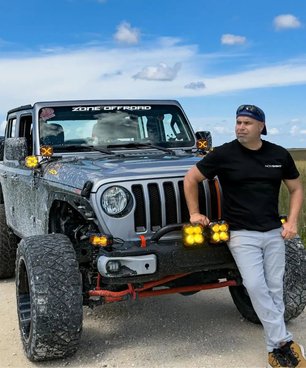 Man standing next to a modified Jeep Wrangler with additional lights on a clear day.