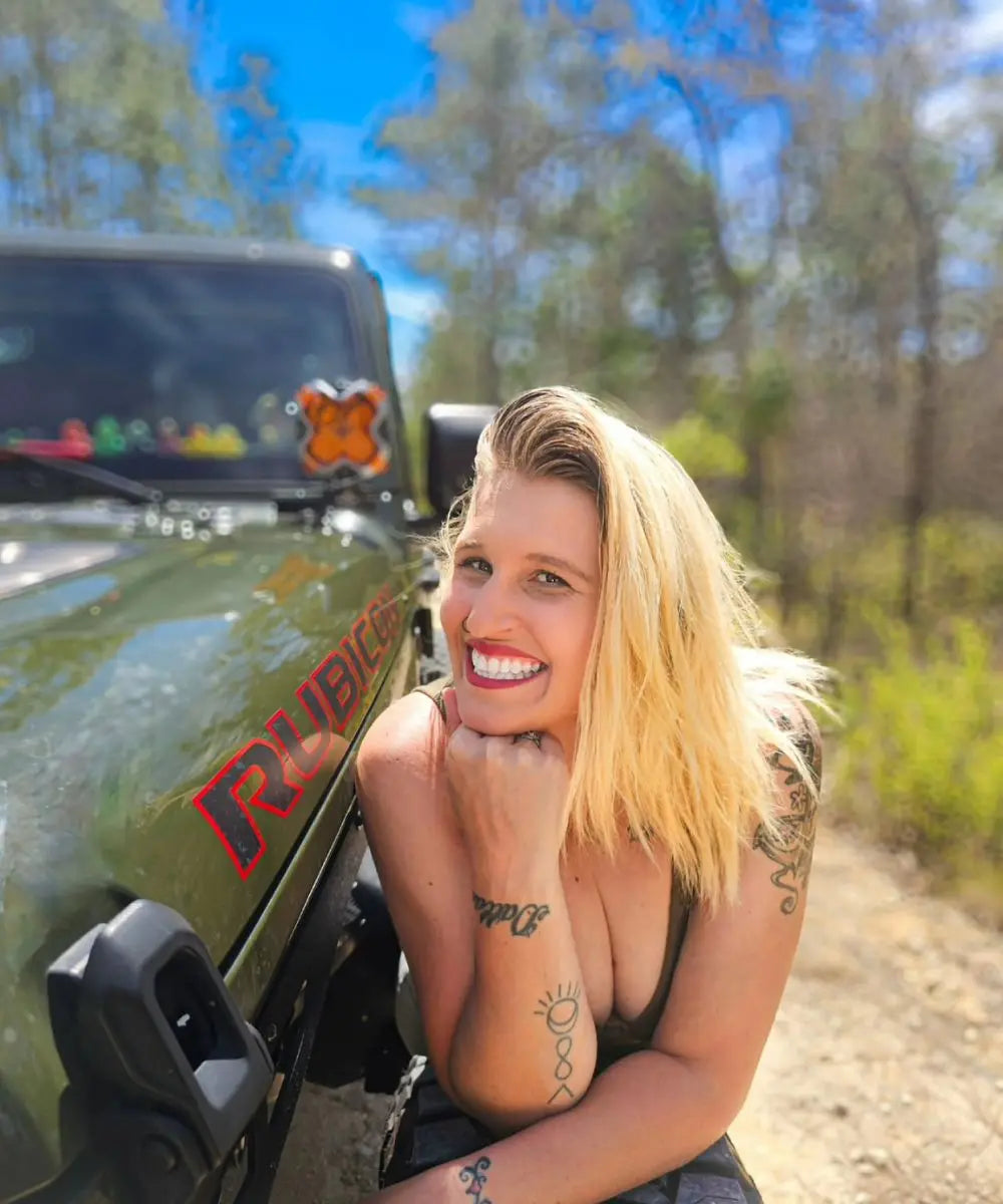 Woman with tattoos leaning against a green Jeep in a forest setting