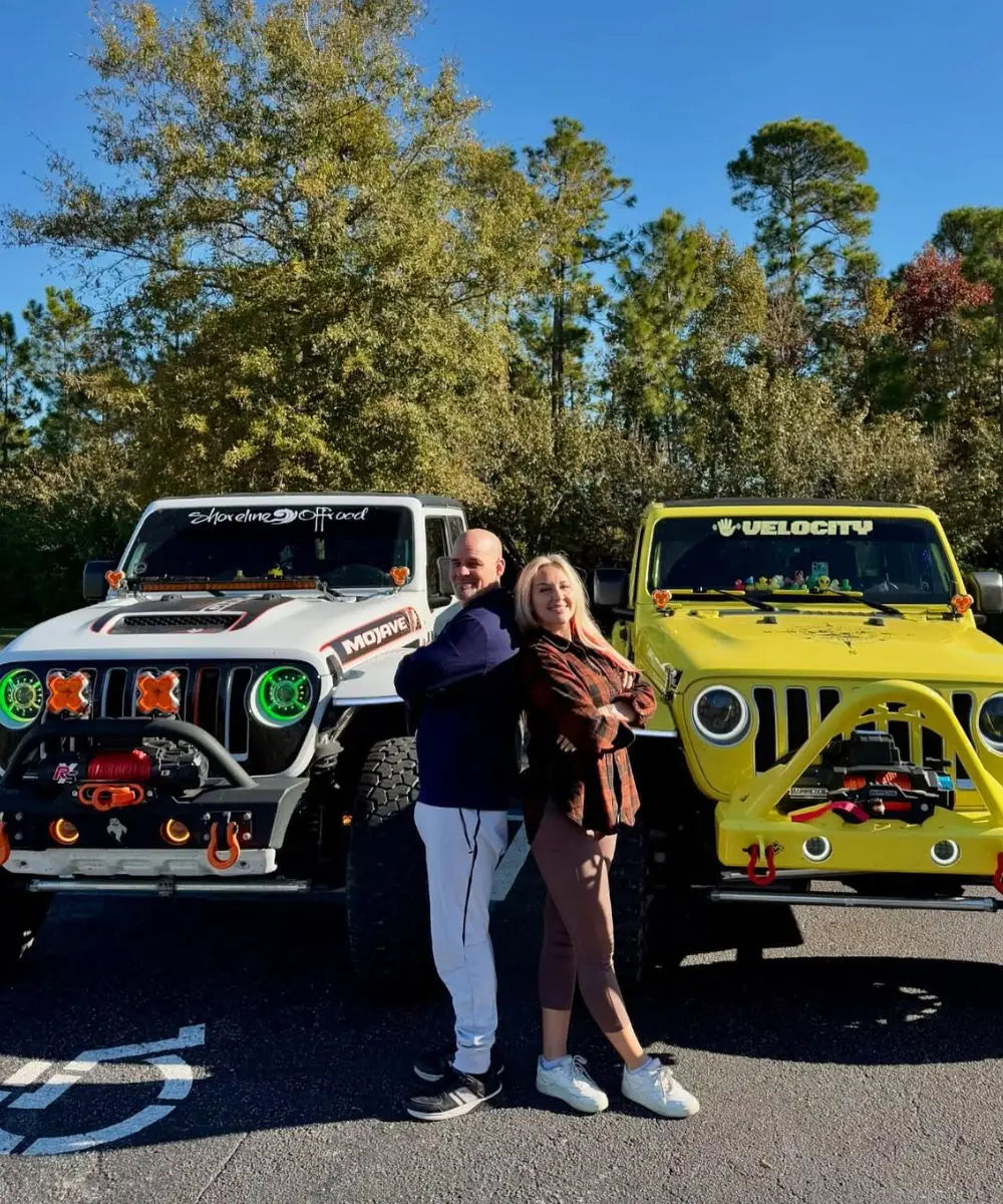 Two people standing between two Jeeps with visible branding in a forested area.