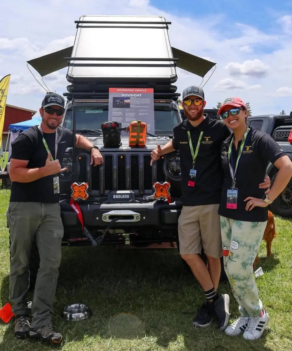 Three people posing in front of a vehicle with outdoor gear at an event.