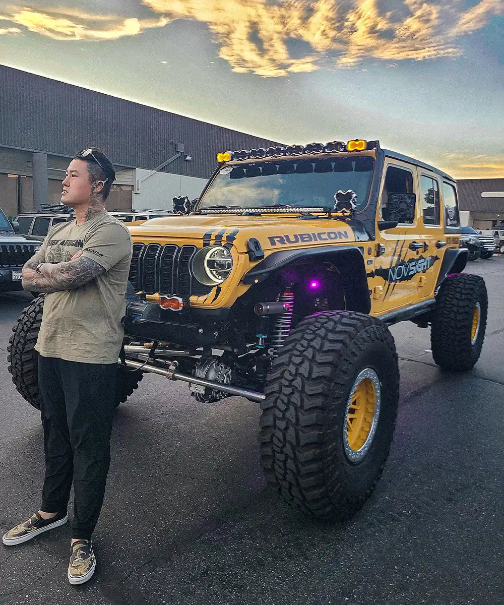 Man standing next to a yellow Jeep Wrangler with large off-road tires in an industrial area.